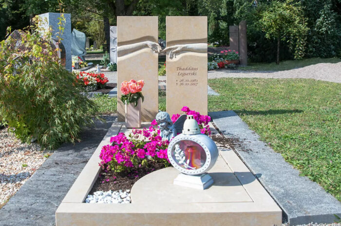 Ein geteiltes Grabmal mit berührenden Händen aus beigem Marmor  umgeben von einer Grabeinfassung und einem Bepflanzungsstreifen als Beispiel für eine Sommergrabgestaltung.