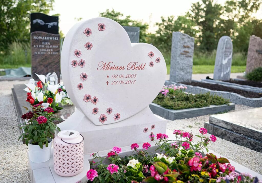 Grabstein als Herz mit Einfassung und Sockel aus hellem Kalkstein mit gravierten Blüten - bunte Sommerbepflanzung mit Blumen