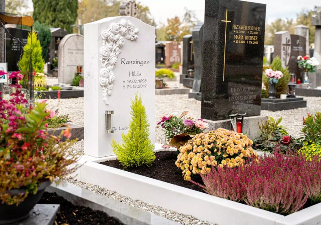 Ein klassischer Grabstein mit einem Rosenornament, umgeben von einer Grabeinfassung aus weißem Marmor sowie Grabgestaltung mit verschiedenen blühenden Pflanzen.