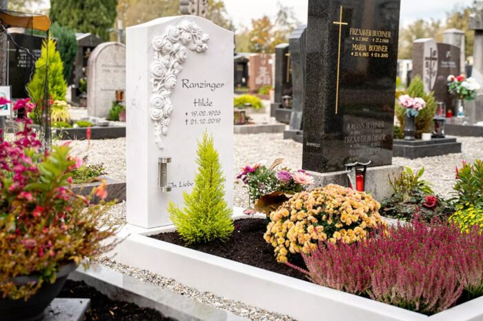 Ein klassischer Grabstein mit einem Rosenornament  umgeben von einer Grabeinfassung aus weißem Marmor sowie Grabgestaltung mit verschiedenen blühenden Pflanzen.