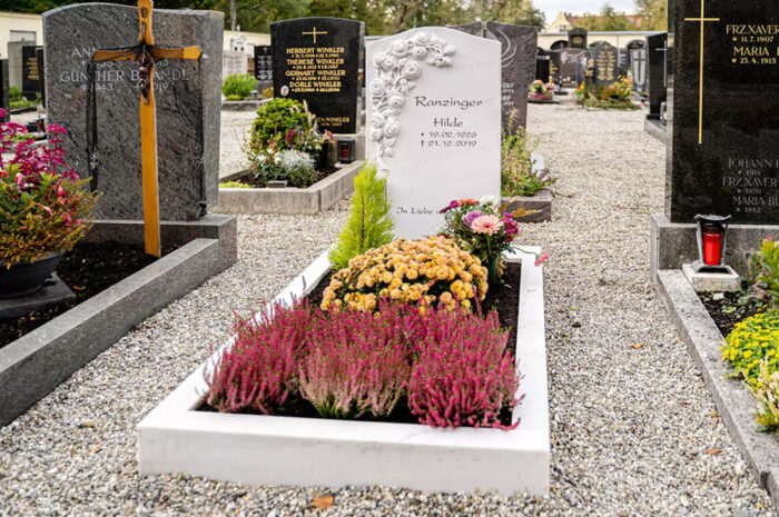 Ein klassischer Grabstein mit einem Rosenornament  umgeben von einer Grabeinfassung aus weißem Marmor sowie Grabgestaltung mit verschiedenen blühenden Pflanzen.